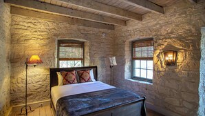 A stone-walled bedroom with a wooden bed, two windows with blinds, a floor lamp, and a wall-mounted light fixture. - Charming Hill Country Getaway in Texas (Fredericksburg)