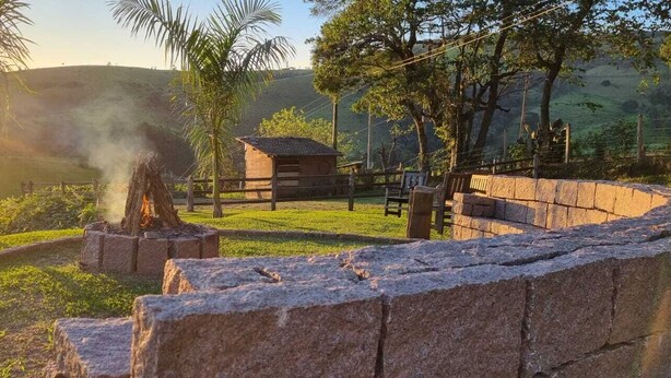 A rustic outdoor setting with a stone wall, a small wooden structure, and a fire burning in a stone-built fireplace. - Pousada Cafuné  (Socorro)