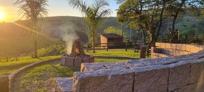 A rustic outdoor setting with a stone wall, a small wooden structure, and a fire burning in a stone-built fireplace. - Pousada Cafuné  (Socorro)
