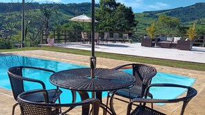 An outdoor pool area with wicker furniture and a large umbrella, surrounded by a scenic view of hills and trees. - Pousada Cafuné  (Socorro)