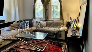 Un salón con sofá, una mesa de centro de cristal, un tapete estampado, una mesita auxiliar con lámpara y una ventana con cortinas verdes.