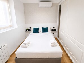 A hotel room with a bed, bedside tables, a radiator, a window, and an air conditioning unit.