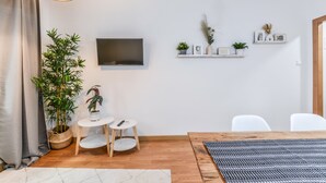 Una sala de estar moderna con una mesa de madera, sillas blancas, un televisor montado en la pared y una planta en maceta.