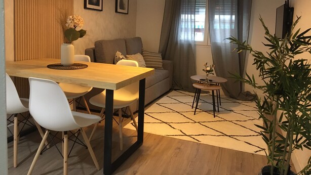 Una sala de estar moderna con una mesa de comedor de madera, sillas blancas, un sofá gris y una mesita auxiliar con un florero.