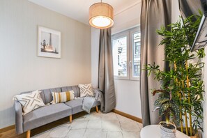 Un salon moderne avec un canapé gris, un tapis à motifs, une grande plante en pot et un tableau encadré représentant la Tour Eiffel.
