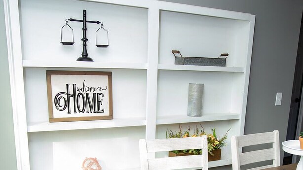 Un comedor con una mesa de madera, sillas blancas, mantel a cuadros, y un estante con un letrero que dice 'Welcome Home' y una balanza.