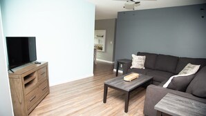 Una sala de estar moderna con un televisor de pantalla plana sobre un mueble de madera, un sofá gris con almohadones decorativos, una mesa de centro y piso de madera.