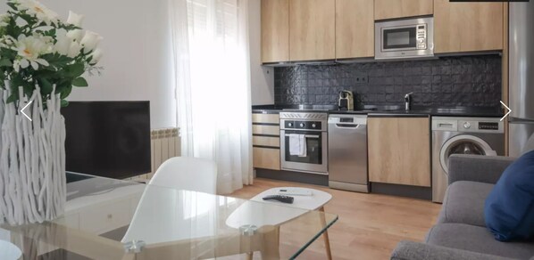 A modern kitchen with stainless steel appliances, a dining area with a glass table, and a living area with a sofa.