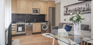 A compact kitchen with modern appliances, a dining area with a glass table, and a living room with a sofa and a cityscape painting. - Collection Madrid Rio Exterior 3 Personas (Madrid)