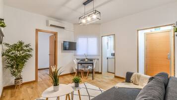 A modern living room with a grey sofa, a small dining table with chairs, a TV mounted on the wall, and a wooden door.