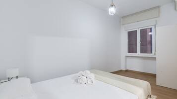 A minimalist bedroom with a single bed, a bedside table with a lamp, a window with blinds, and a hanging light fixture.
