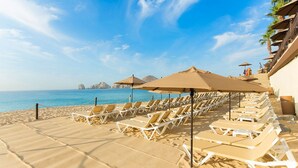 A beachside resort with rows of sun loungers and umbrellas, a clear view of the sea, and a rocky coastline in the background.