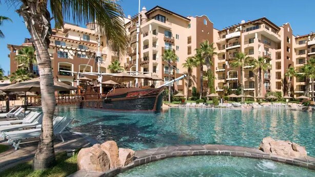 A resort pool with lounge chairs, palm trees, and a large building with multiple balconies.