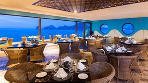 A restaurant with tables set for dining, large windows offering a view of the sea, and a ceiling with a geometric pattern.
