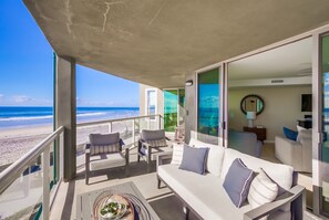Un balcón con vista a la playa, amueblado con un sofá y sillones, y una zona de comedor con mesa y sillas.