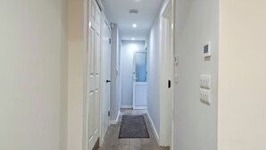 A narrow hallway with white walls, wooden flooring, and a dark rug leading to a door at the end.