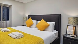 A modern bedroom with a large bed, two yellow pillows, a white blanket, and a yellow throw.