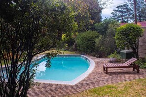 A rectangular swimming pool with a brick deck surrounding it, a wooden lounge chair, and lush greenery in the background.