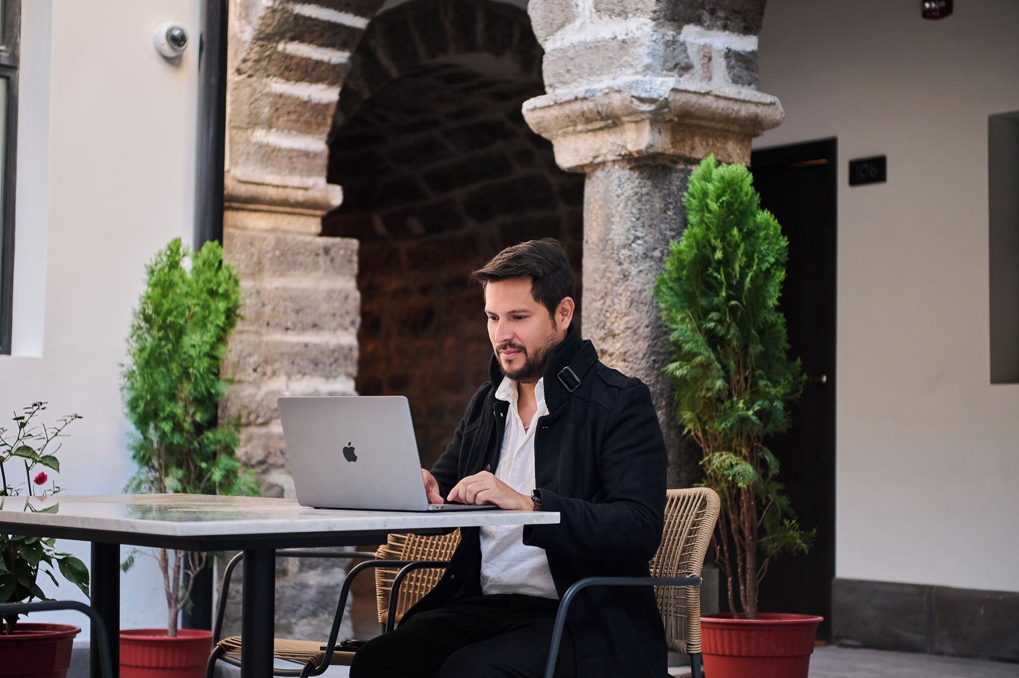 Una persona usando una computadora portátil en una mesa al aire libre con plantas en macetas, frente a un arco de piedra.