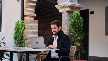 Una persona usando una computadora portátil en una mesa al aire libre con plantas en macetas, frente a un arco de piedra.