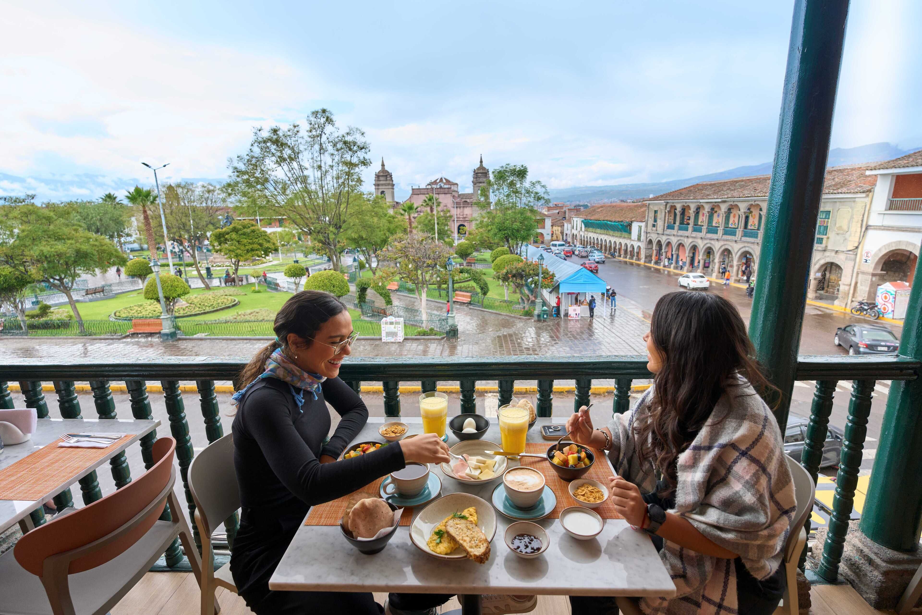 Un balcón con una mesa lista para el desayuno, con vistas a un paisaje urbano que incluye edificios históricos y un parque.