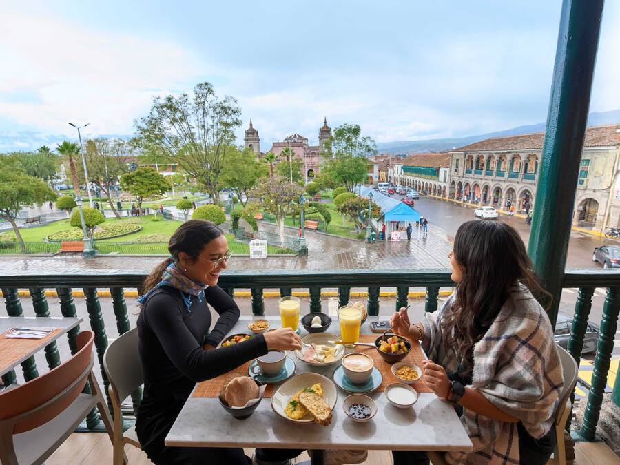 Un balcón con una mesa lista para el desayuno, con vistas a un paisaje urbano que incluye edificios históricos y un parque.