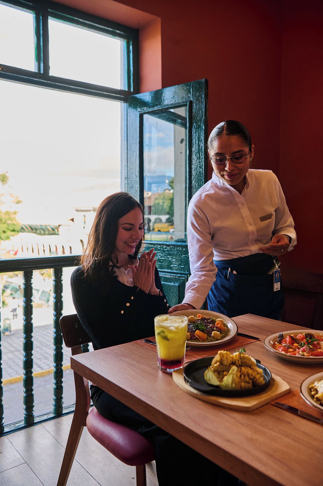 Un interior de restaurante con una mesa de madera, vistas al exterior y una persona de pie junto a la mesa.