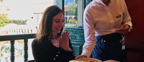 Un interior de restaurante con una mesa de madera, vistas al exterior y una persona de pie junto a la mesa.
