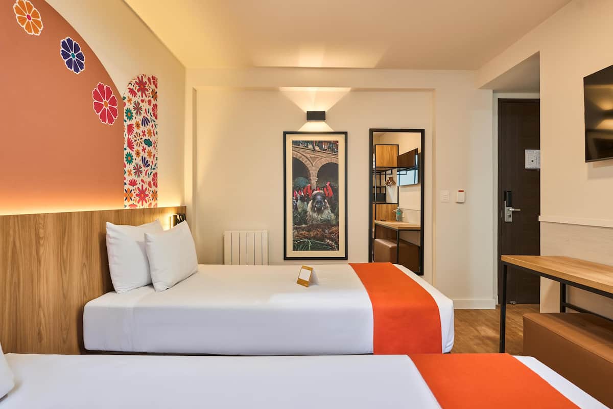 Ein Hotelzimmer mit zwei Betten, einem hölzernen Kopfteil, einem an der Wand montierten Fernseher und einem gerahmten Bild.