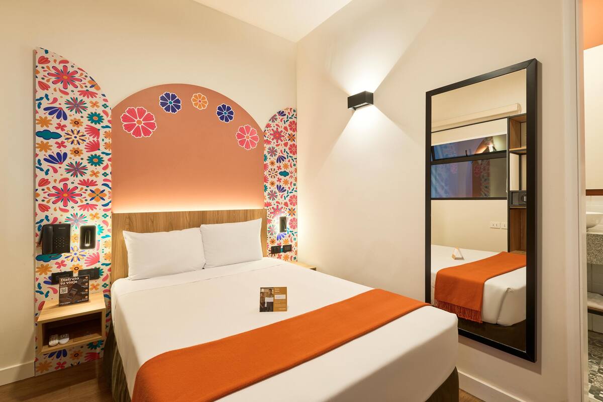 Ein Hotelzimmer mit einem großen Bett, einem Nachttisch mit Telefon und Broschüre, einem Spiegel und einer Wand mit floralem Muster.