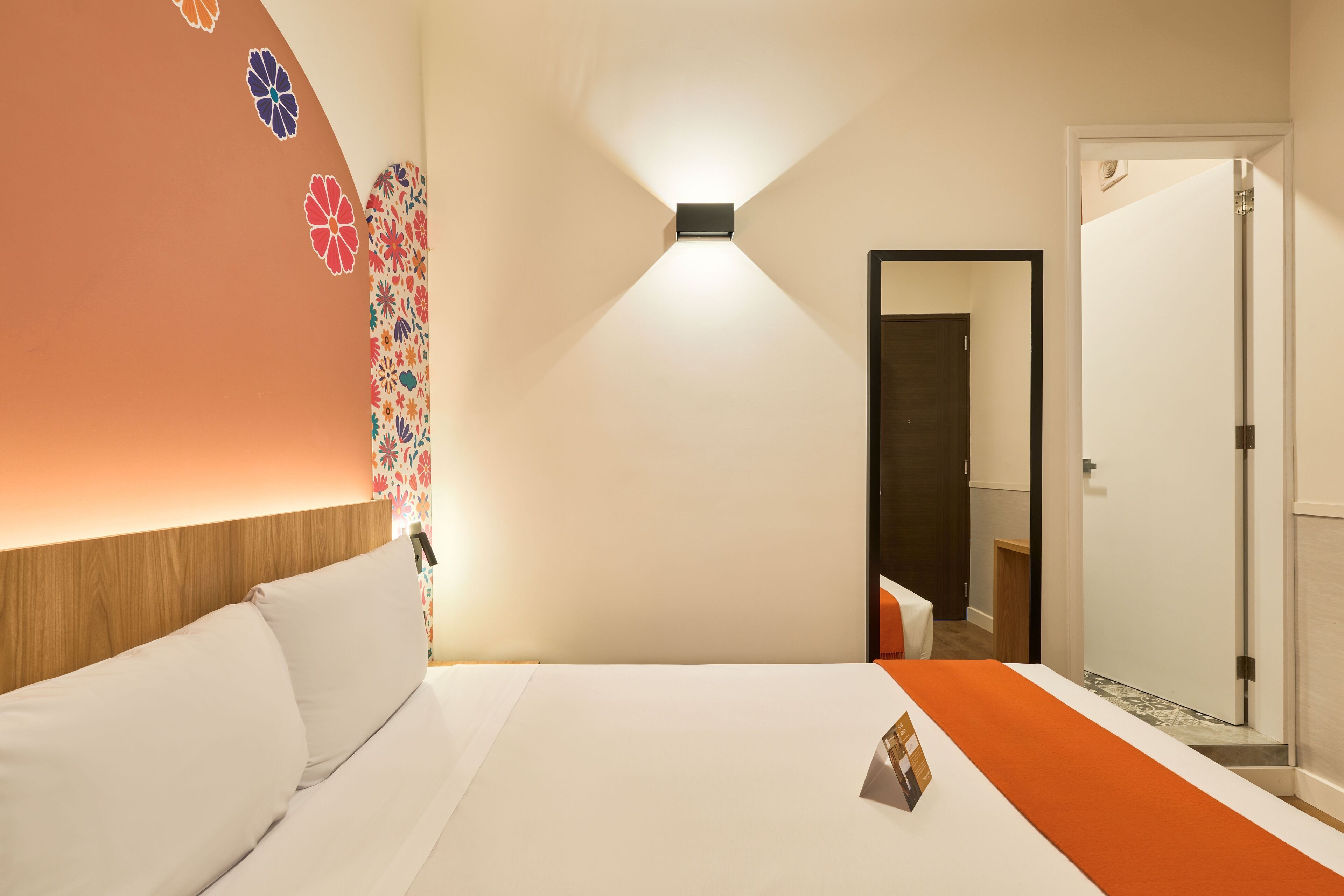 Quarto de hotel com cama, mesa de cabeceira, luminária na parede e porta para outro cômodo.