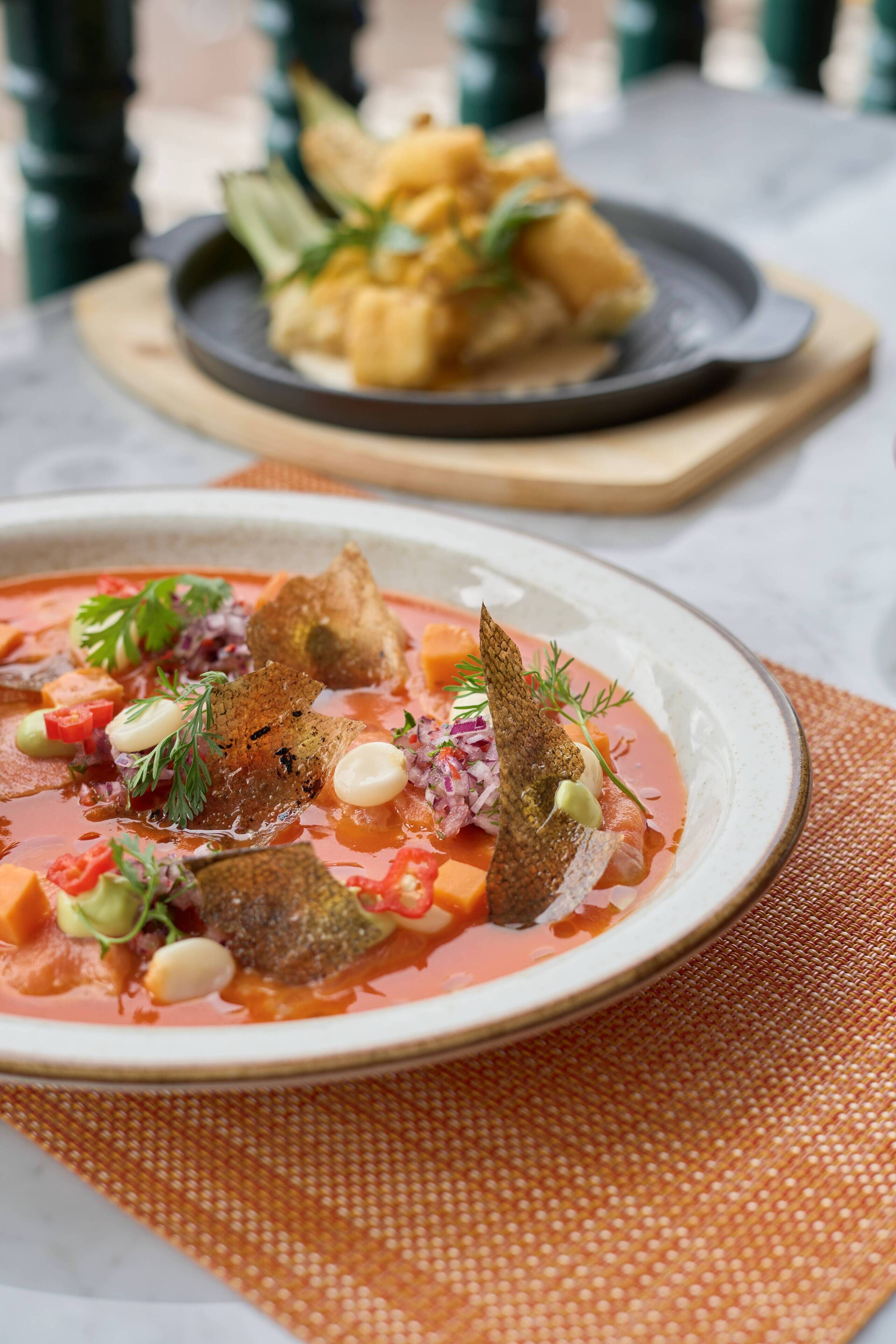 A bowl of soup with various garnishes and a plate of food in the background.