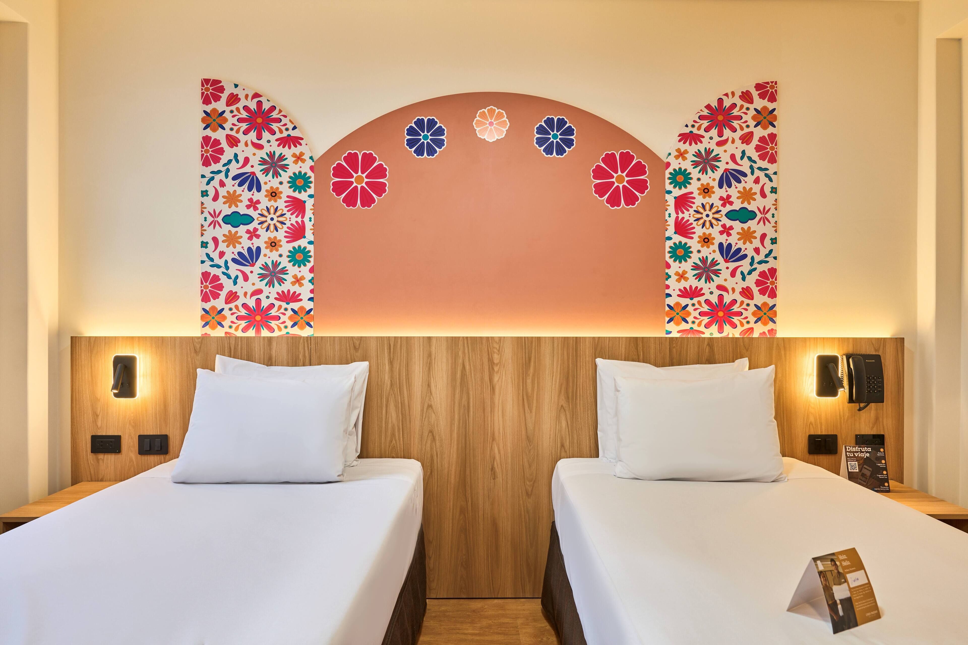 A hotel room with two beds, wooden headboard, wall-mounted phone, and a decorative floral pattern on the wall.