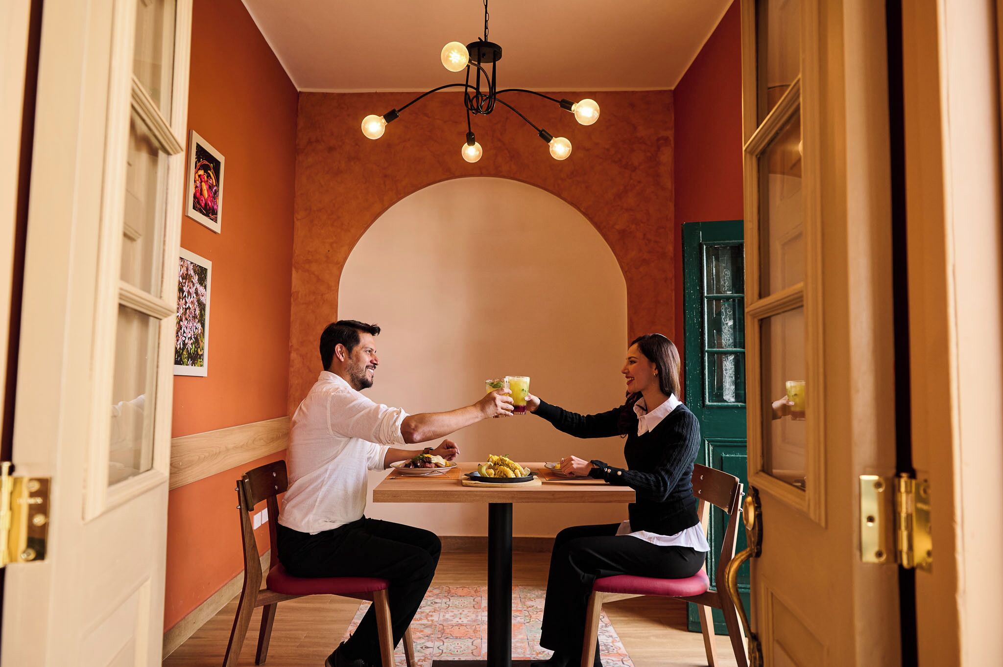 Una pareja brindando en una mesa, con una araña colgando del techo, en una habitación con un arco y una puerta verde.