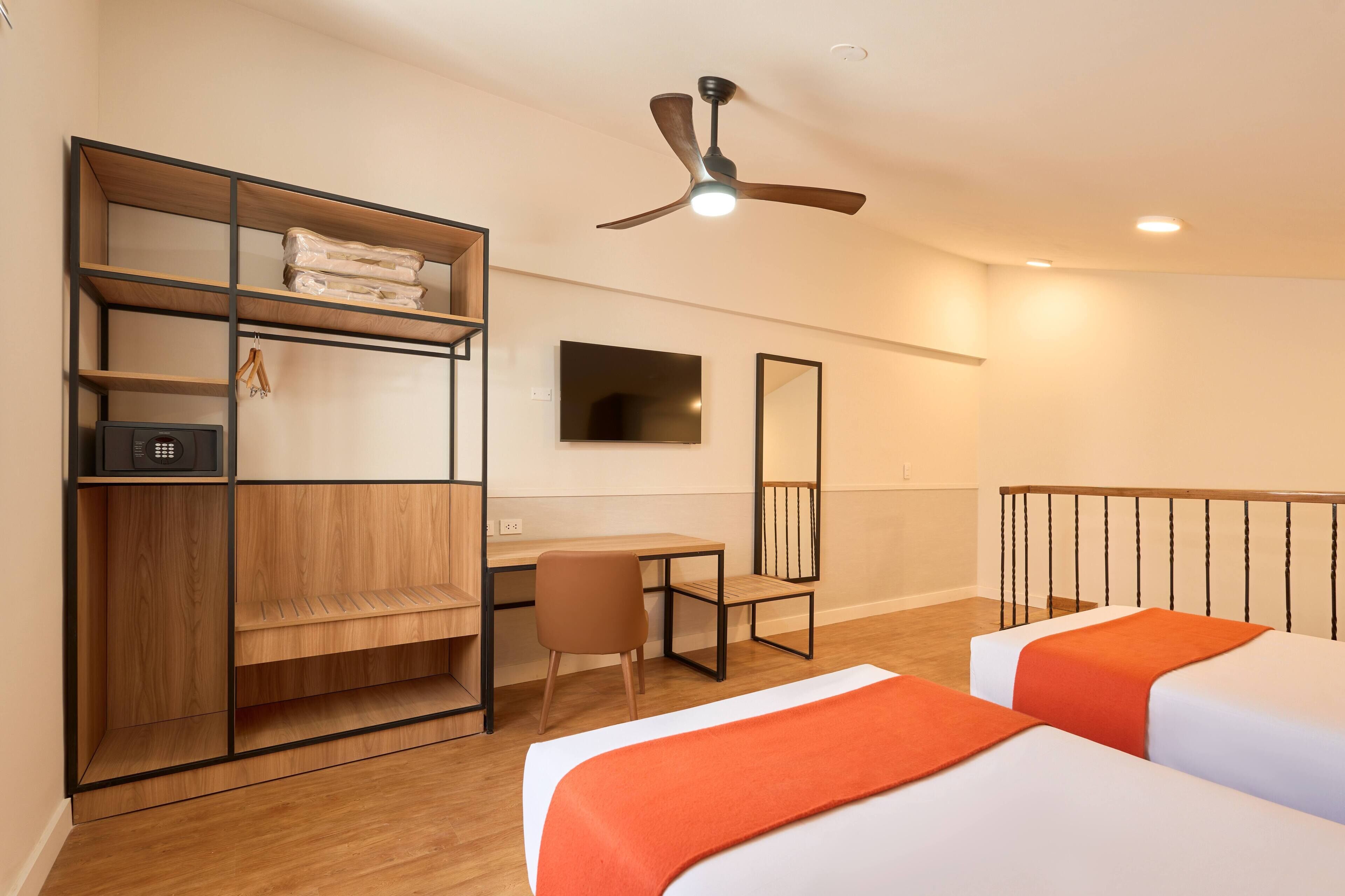 Habitación de hotel con cama, escritorio, silla, armario, ventilador de techo, televisión y barandilla de balcón.