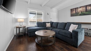 Un salon moderne avec un canapé d’angle, une table basse ronde, un téléviseur fixé au mur et un tableau représentant une forêt.