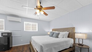 Ein Schlafzimmer mit Bett, Deckenventilator, Klimaanlage, Mikrowelle, Nachttisch mit Lampe und Fenster mit Jalousien.
