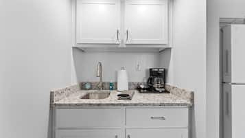 A compact kitchen with white cabinets, a granite countertop, a stainless steel sink, a coffee maker, and a refrigerator.