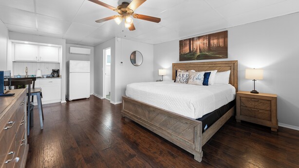 Une chambre avec un lit en bois, une table de chevet, un ventilateur de plafond, un tableau, et un coin cuisine à l’arrière-plan.
