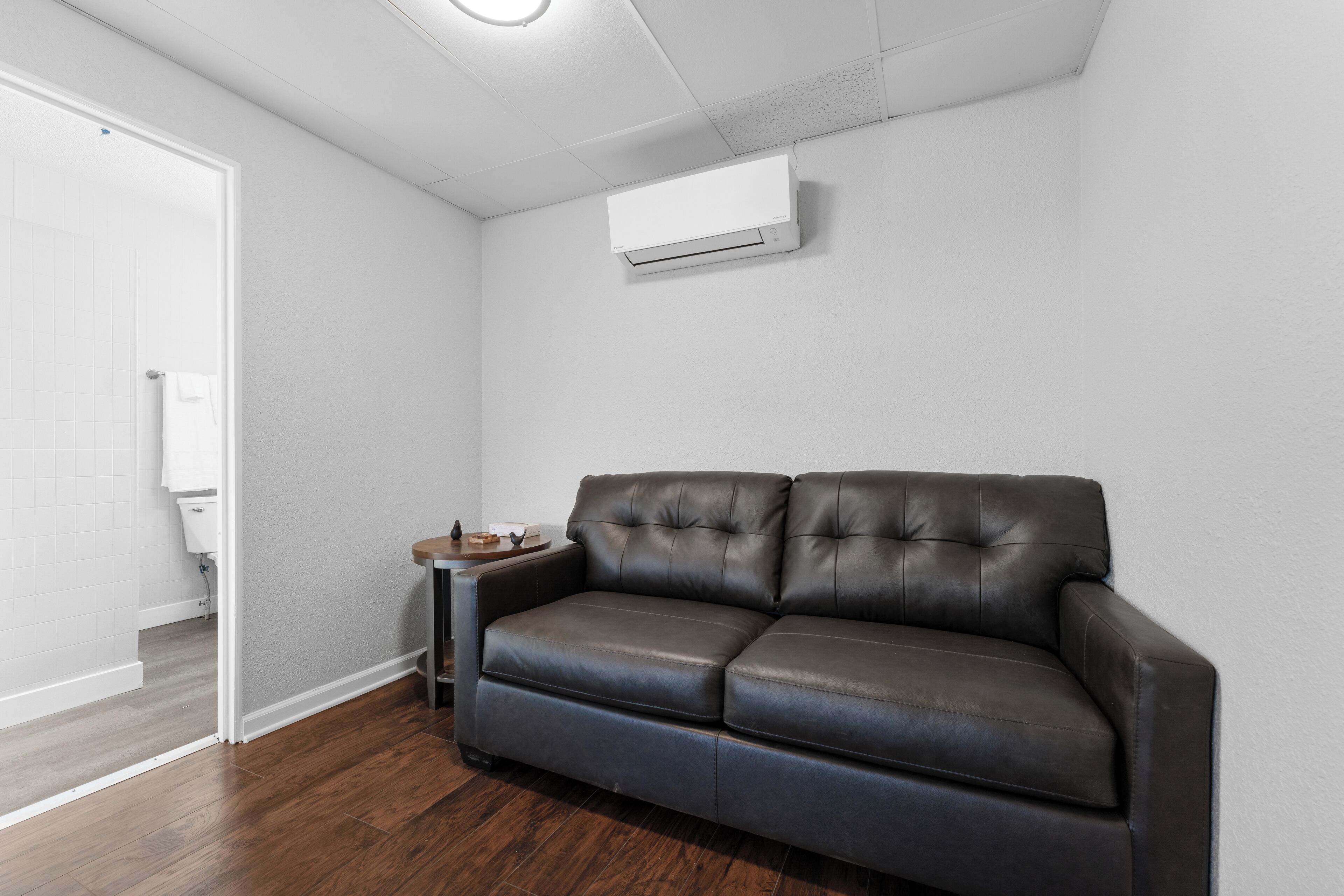 A room with a dark brown leather sofa, a small side table, a white air conditioning unit, and a wooden floor.