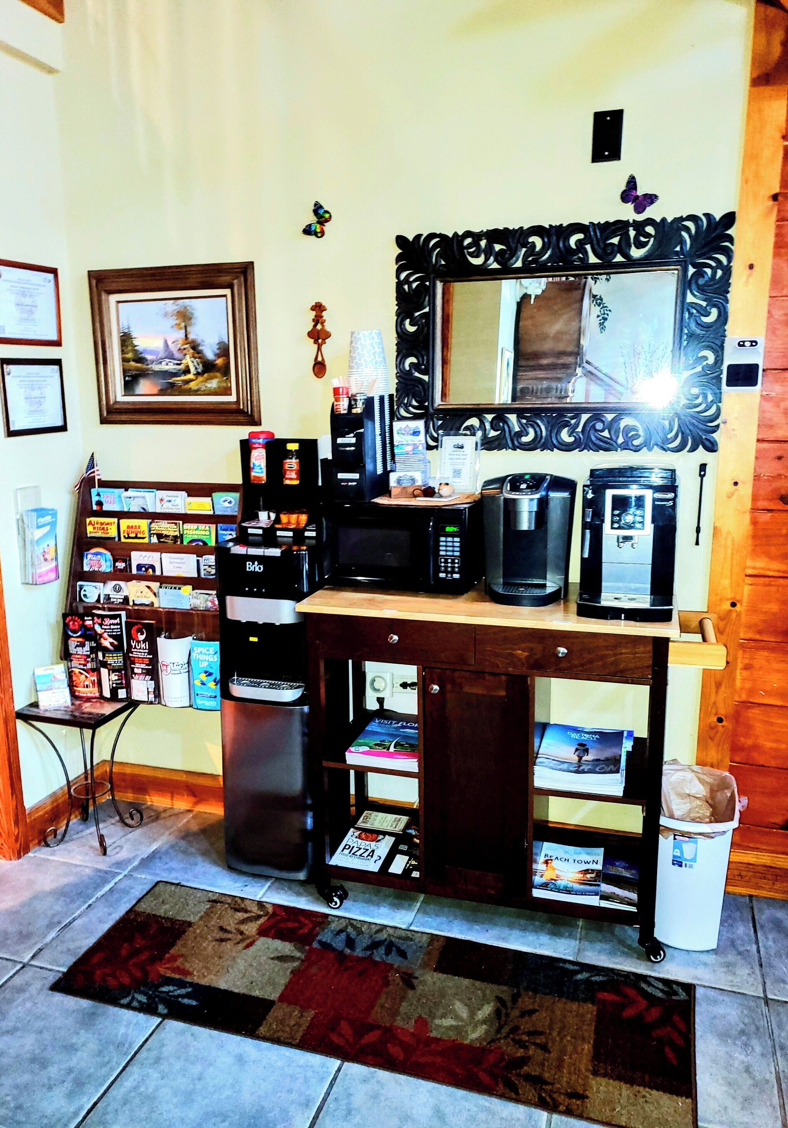 A kitchenette with a coffee maker, microwave, and toaster. There is a small shelf with snacks and a framed picture on the wall.
