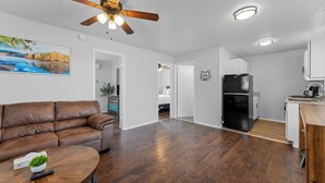 A spacious living area with a brown leather sofa, a round wooden coffee table, and a ceiling fan. A kitchen with white cabinets and a black refrigerator is visible in the background. - Come stay at this modern 2 bedroom suite, located close to Table Rock Lake and Silver Dollar City. (Branson)