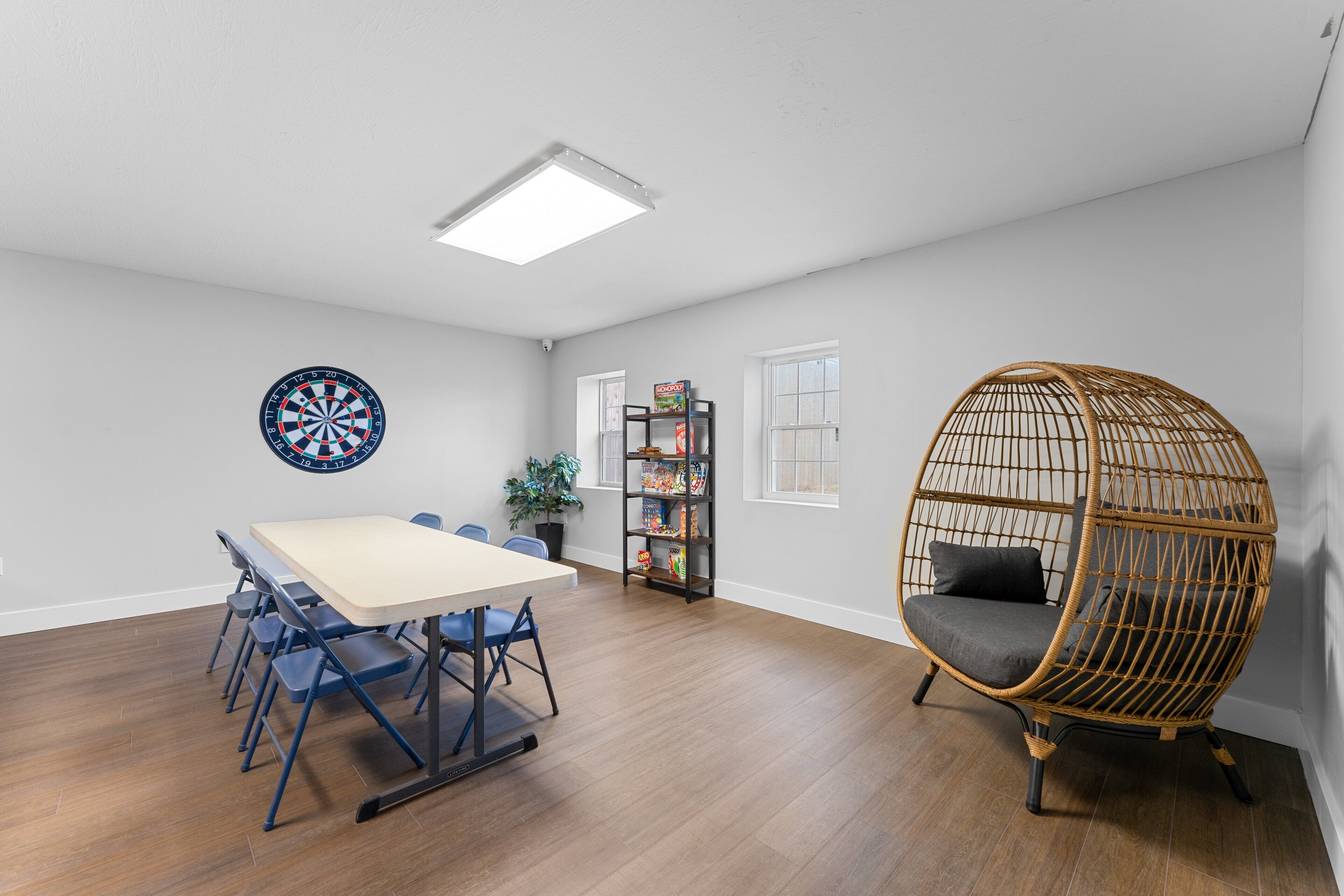 A spacious room with a wooden floor, a round dartboard, a bookshelf, and a wicker chair.