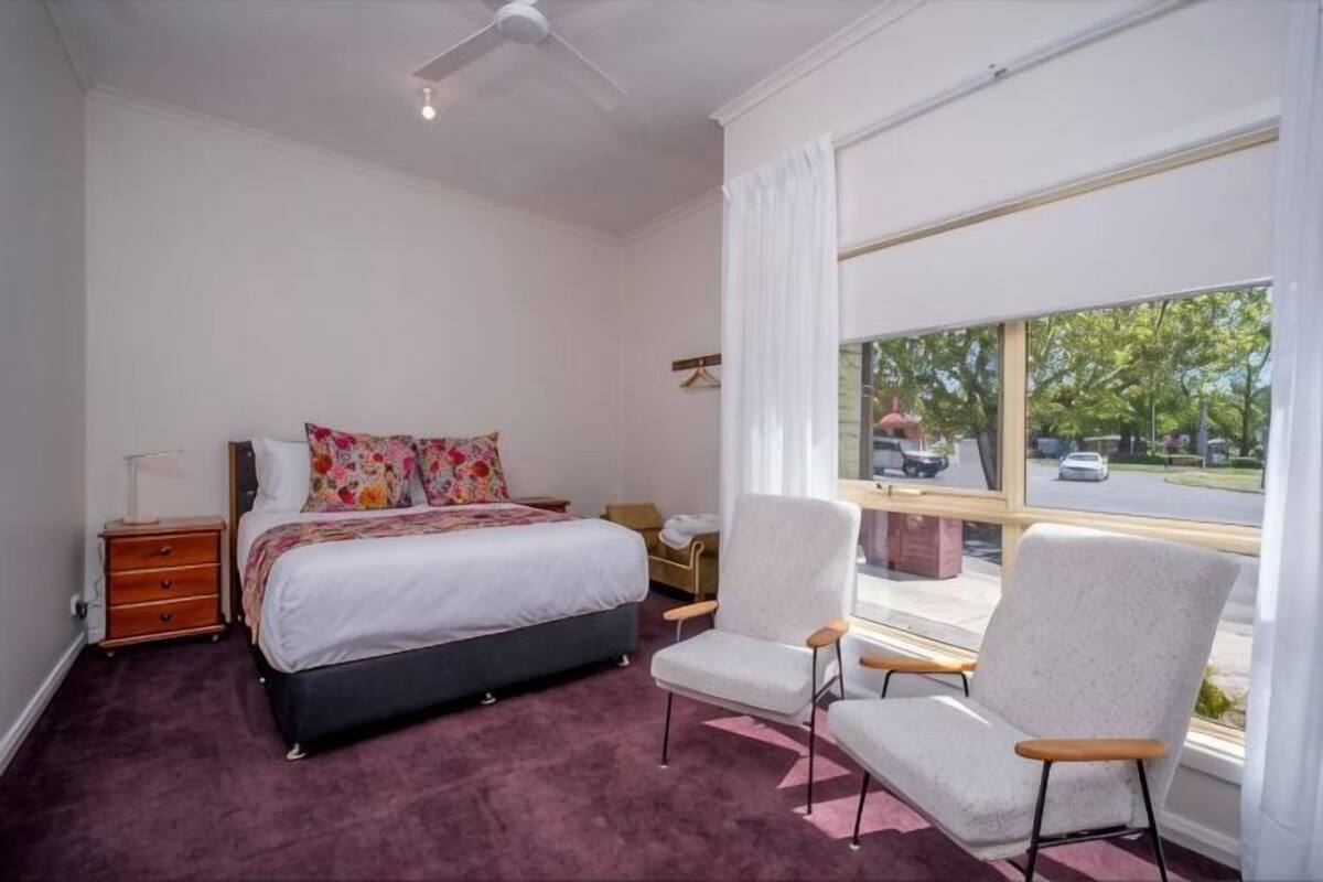 A bedroom with a bed, a chair, a nightstand, a window with a view of the outside, and a ceiling fan.