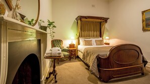 Un dormitorio con una cama con dosel, una chimenea, un sillón verde y una mesita de noche con lámpara.