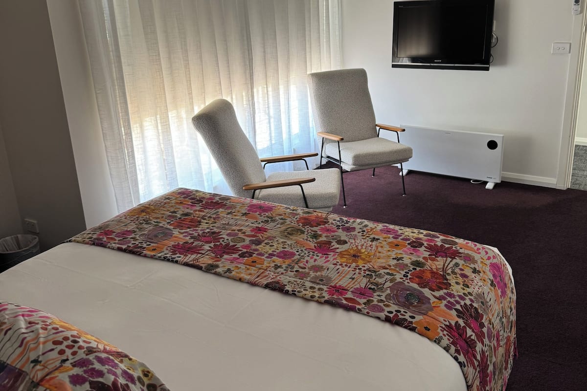 A hotel room with a large bed, two armchairs, a flat-screen TV, and a large window with sheer curtains.