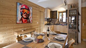 A dining area with a wooden table set for four, a painting on the wall, and a kitchen in the background.