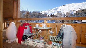 A wooden deck with a table and chairs, a white ceramic rooster, and a view of snow-covered mountains.