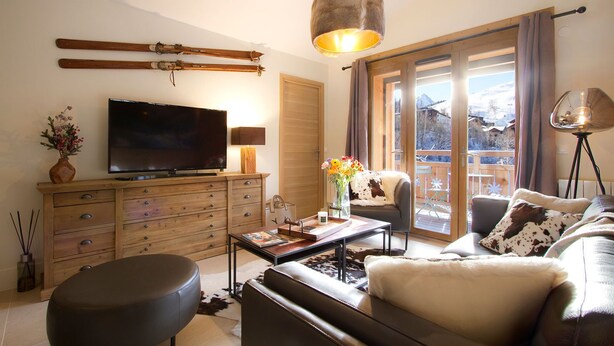A cozy living room with a TV, wooden furniture, a sofa, a coffee table, and a view of a balcony with a wooden deck.