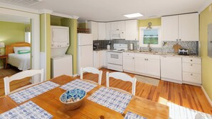 A compact kitchen with white cabinets, a microwave, a stove, and a sink. There is a dining table with chairs and a bowl of fruit.